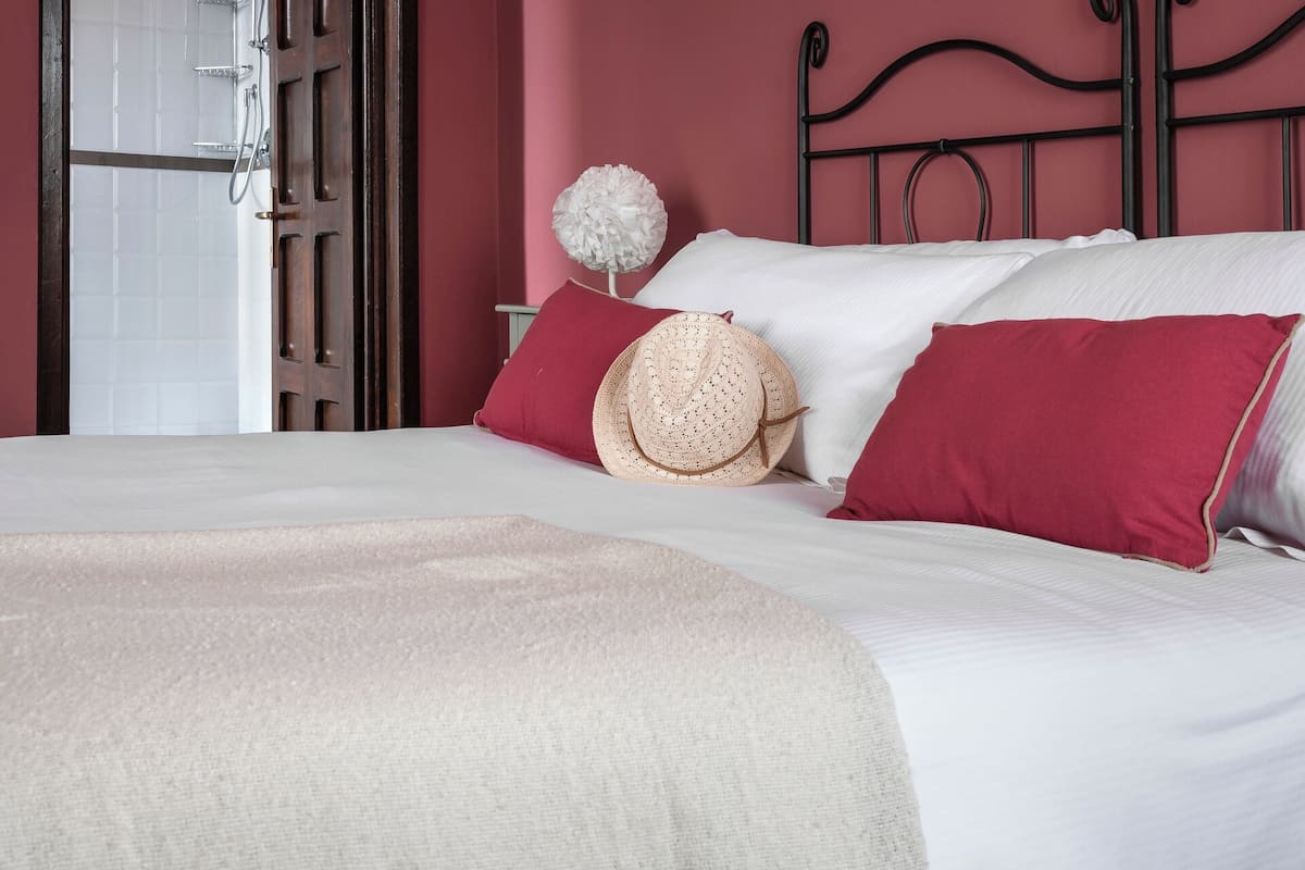 Un lit bien fait, avec des draps blancs et des oreillers rouges, une couverture beige et un chapeau de paille posé sur les oreillers.