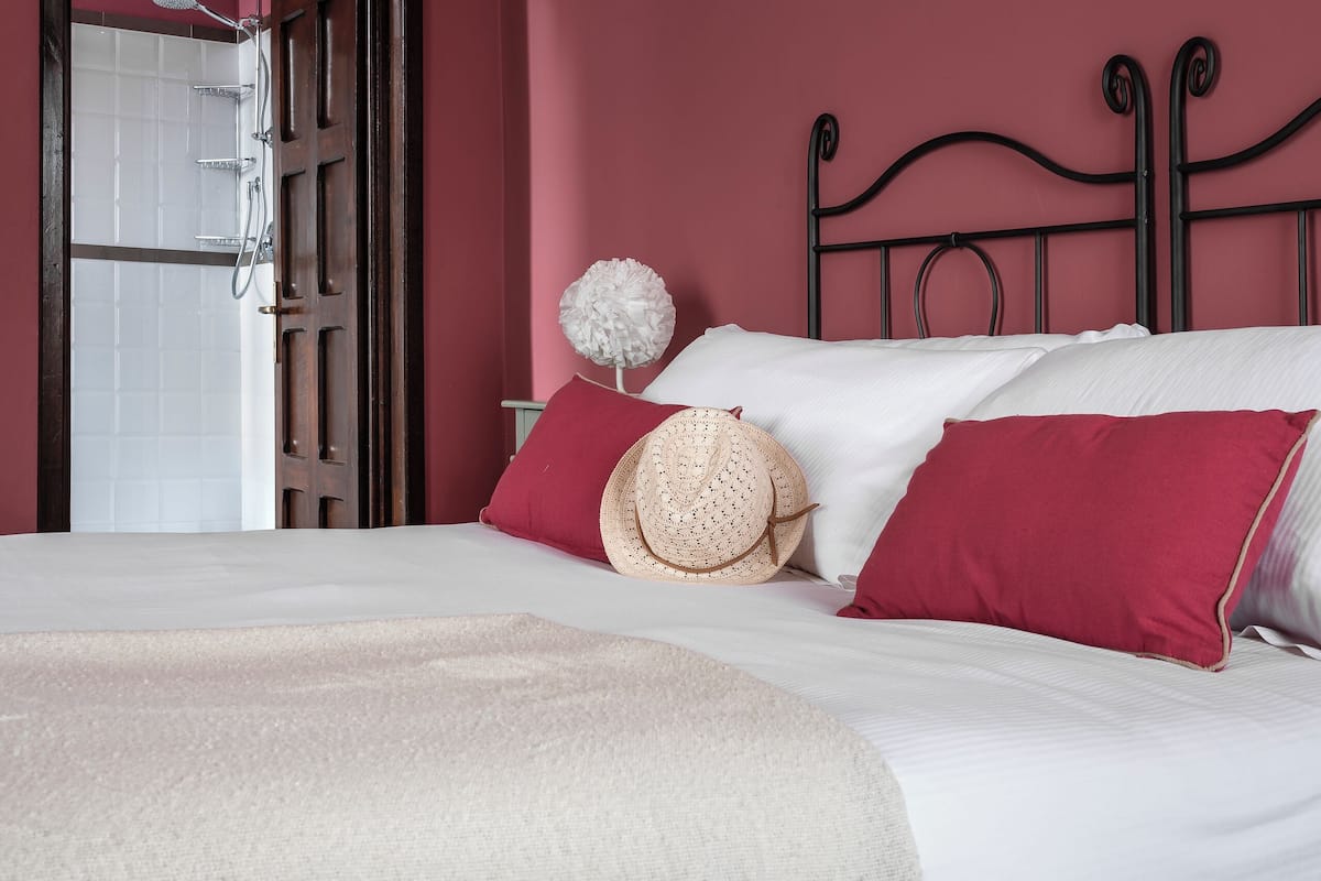 Un lit bien fait, avec du linge de lit blanc et un plaid beige, un coussin rouge et un chapeau de paille posé à côté.
