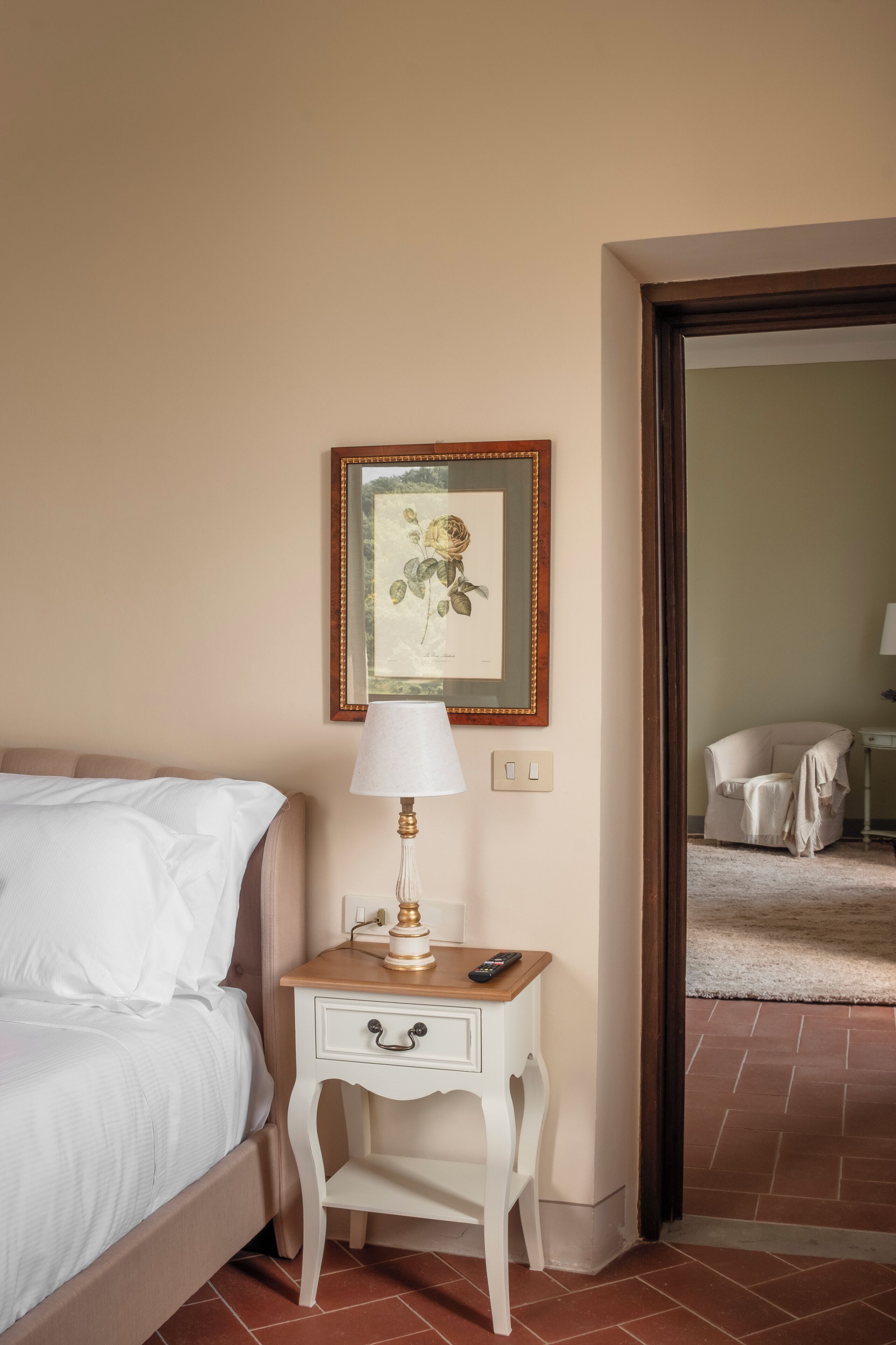 A hotel room with a bed, a bedside table with a lamp, a framed picture on the wall, and a chair in the background.