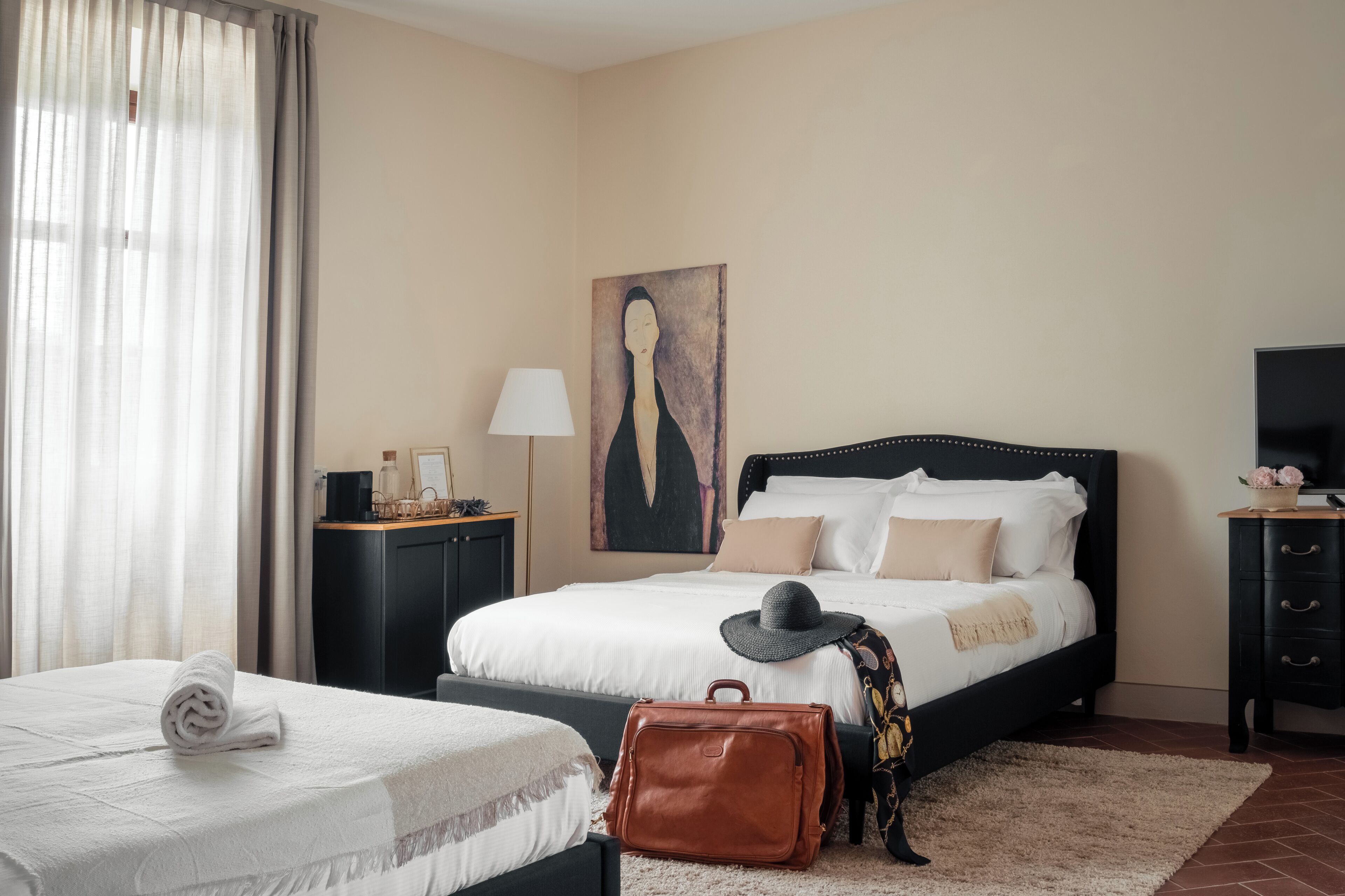 A hotel room with a large bed, a smaller bed, a desk with a lamp, a painting on the wall, and a TV on a stand.