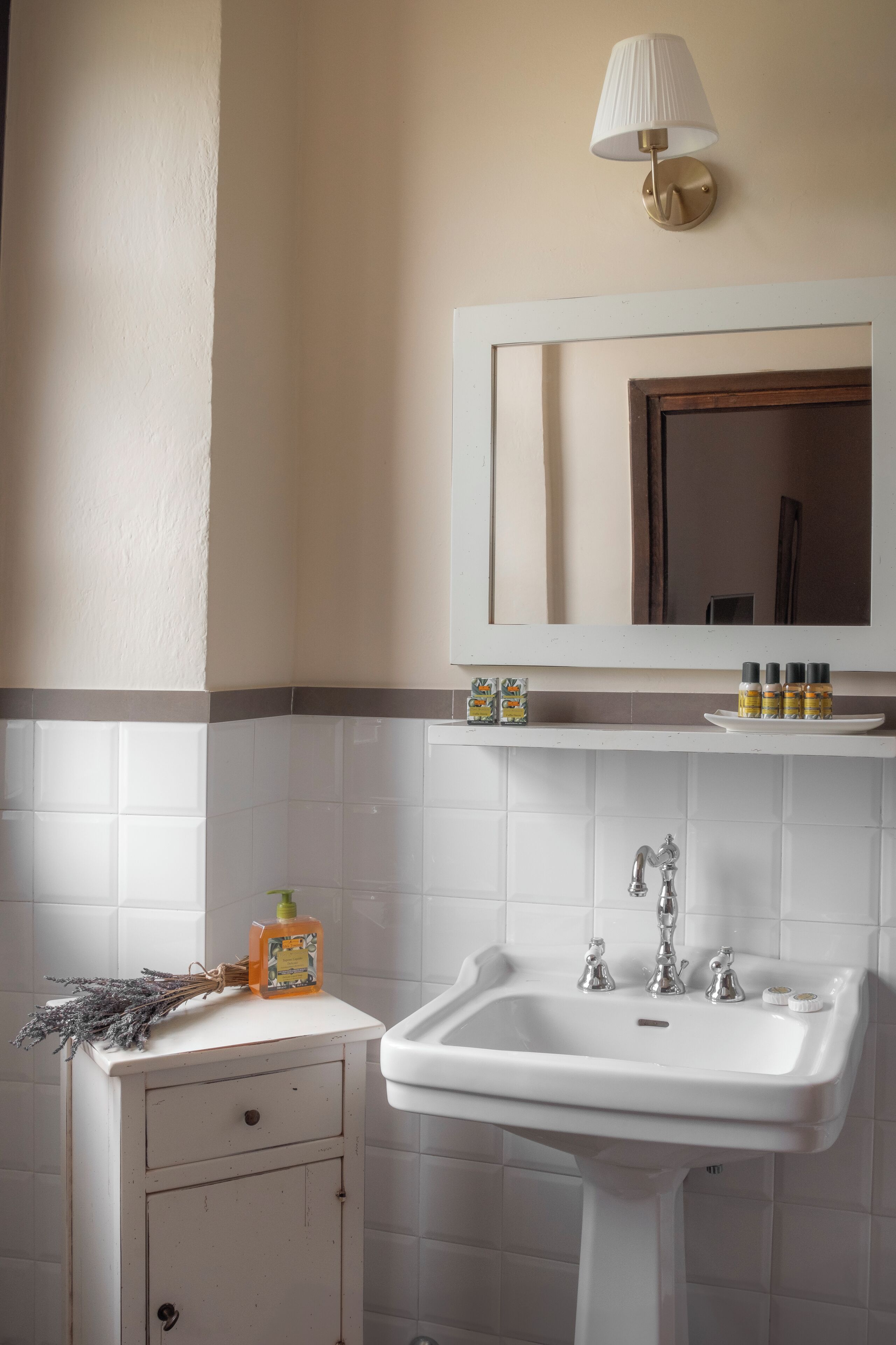 Une salle de bain avec un lavabo blanc, un miroir au-dessus et une lampe fixée au mur. On peut voir des articles de toilette posés sur le bord du lavabo.