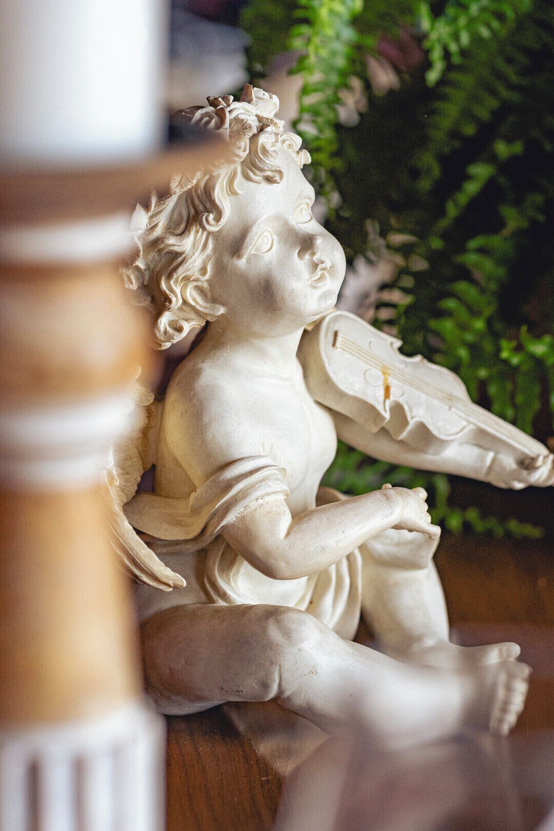 A statue of a cherub holding a violin, set against a backdrop of greenery.
