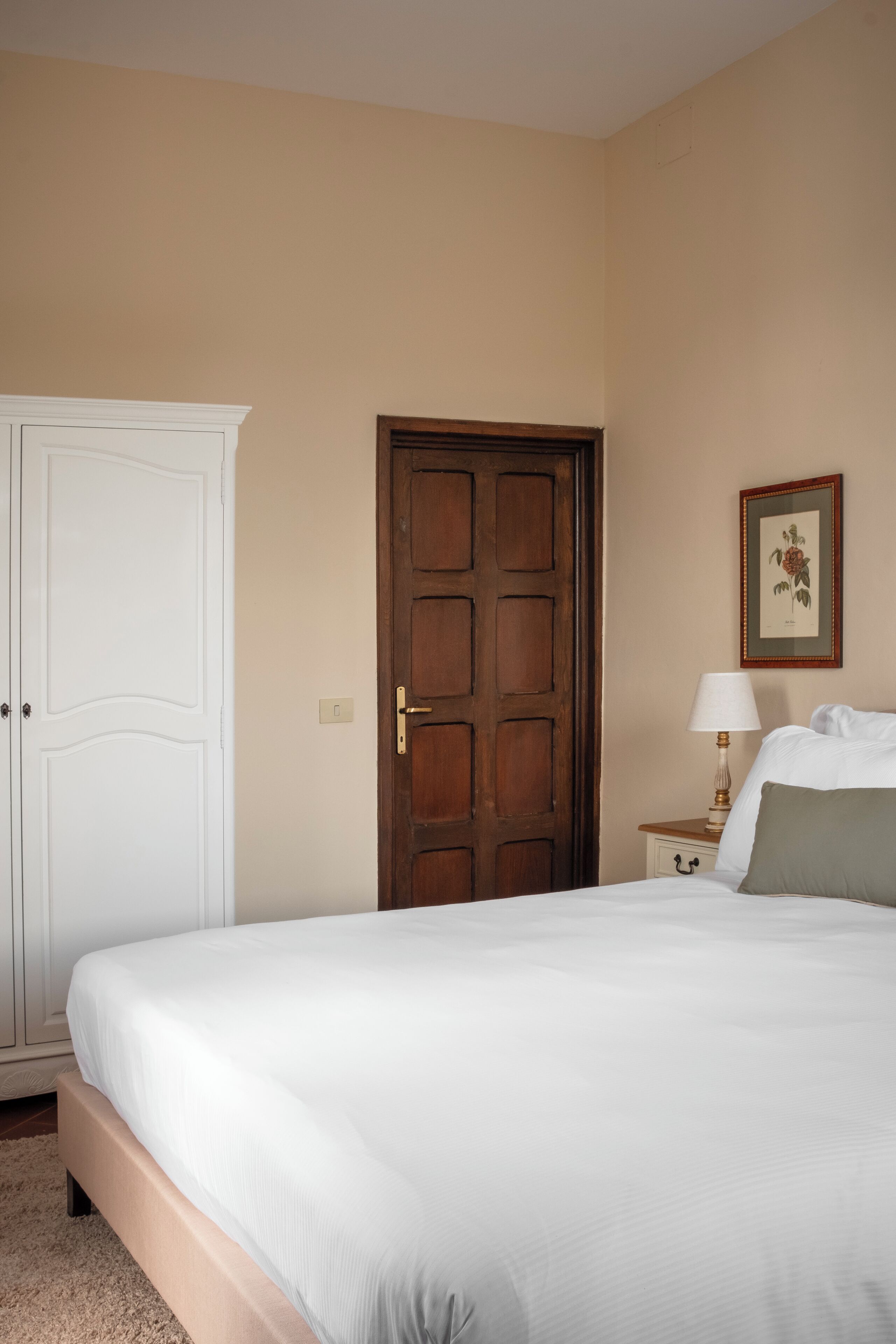 Un dormitorio con cama, armario, puerta, mesita de noche con lámpara y un cuadro en la pared.