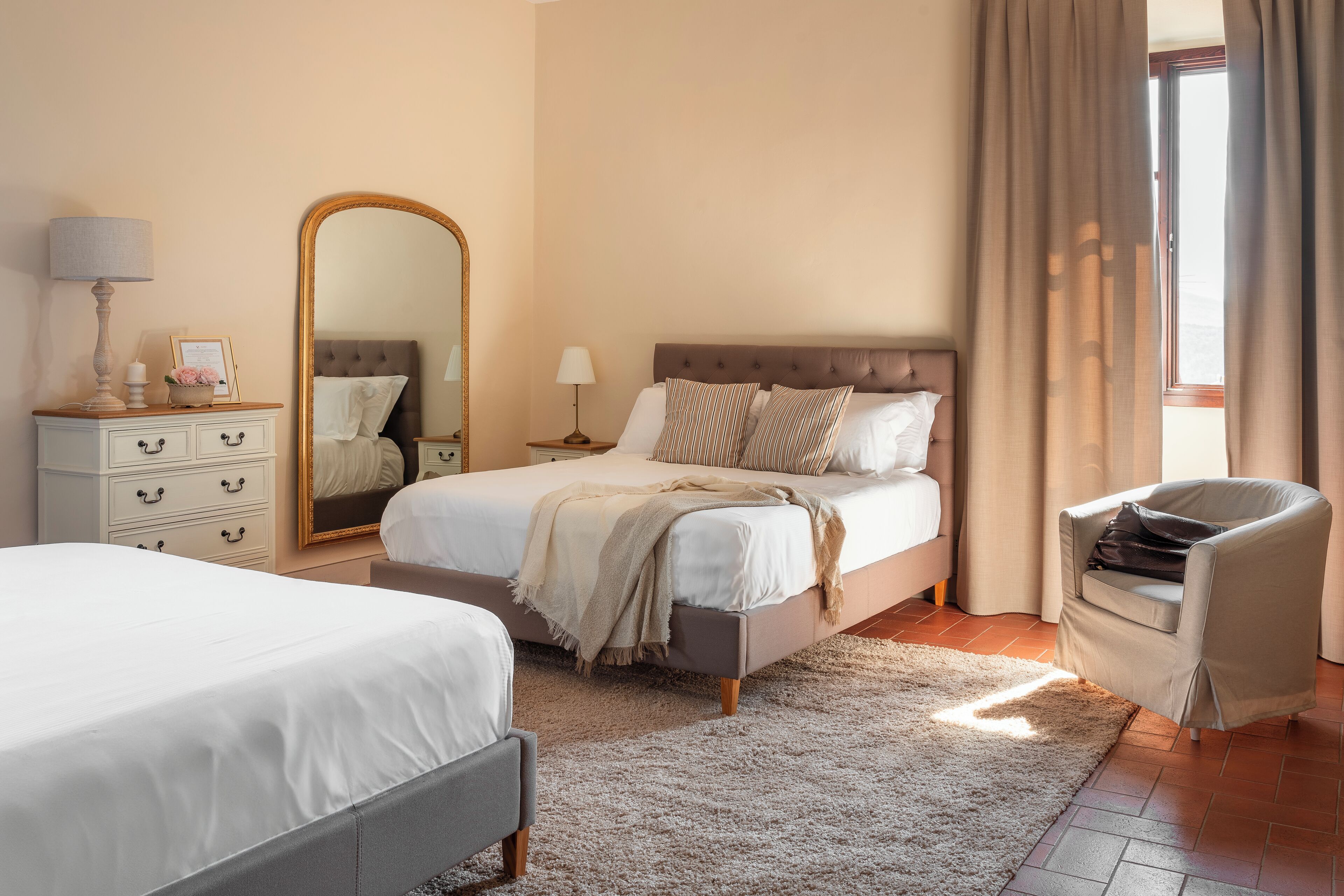 A bedroom with a large bed, a smaller bed, a chair, a dresser, a mirror, and a window with curtains.