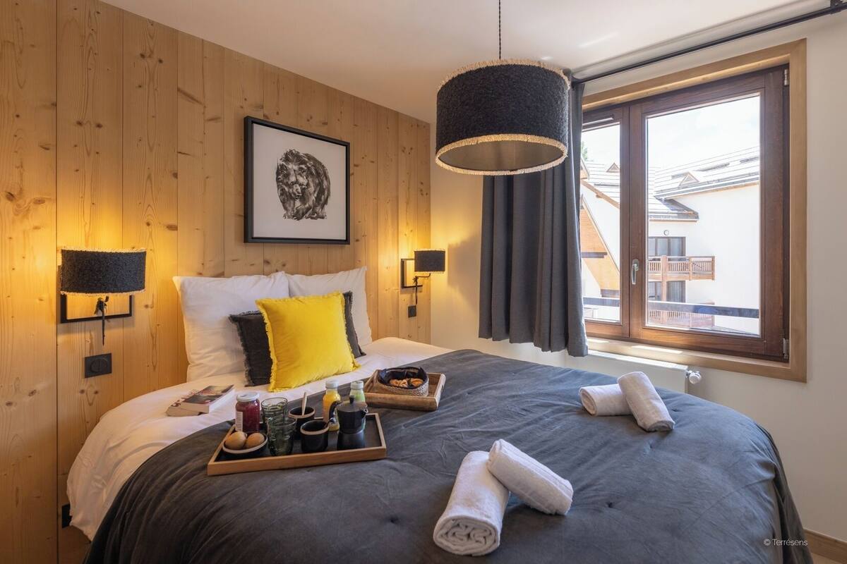 A modern hotel room with a wooden wall, a large bed with grey bedding, a yellow pillow, and a tray of breakfast items.
