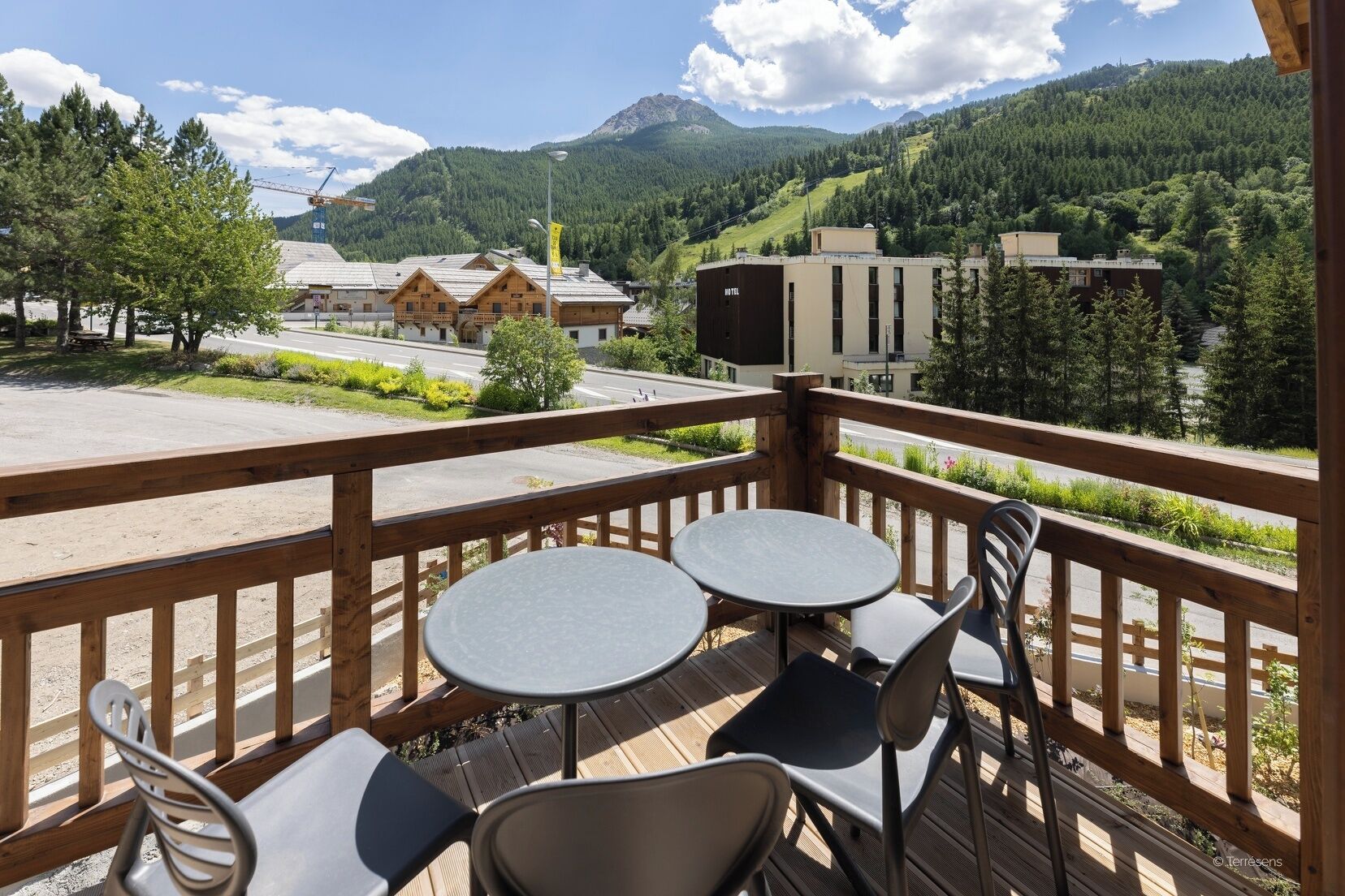 A wooden deck with two round tables and chairs, overlooking a mountainous landscape with buildings in the background.