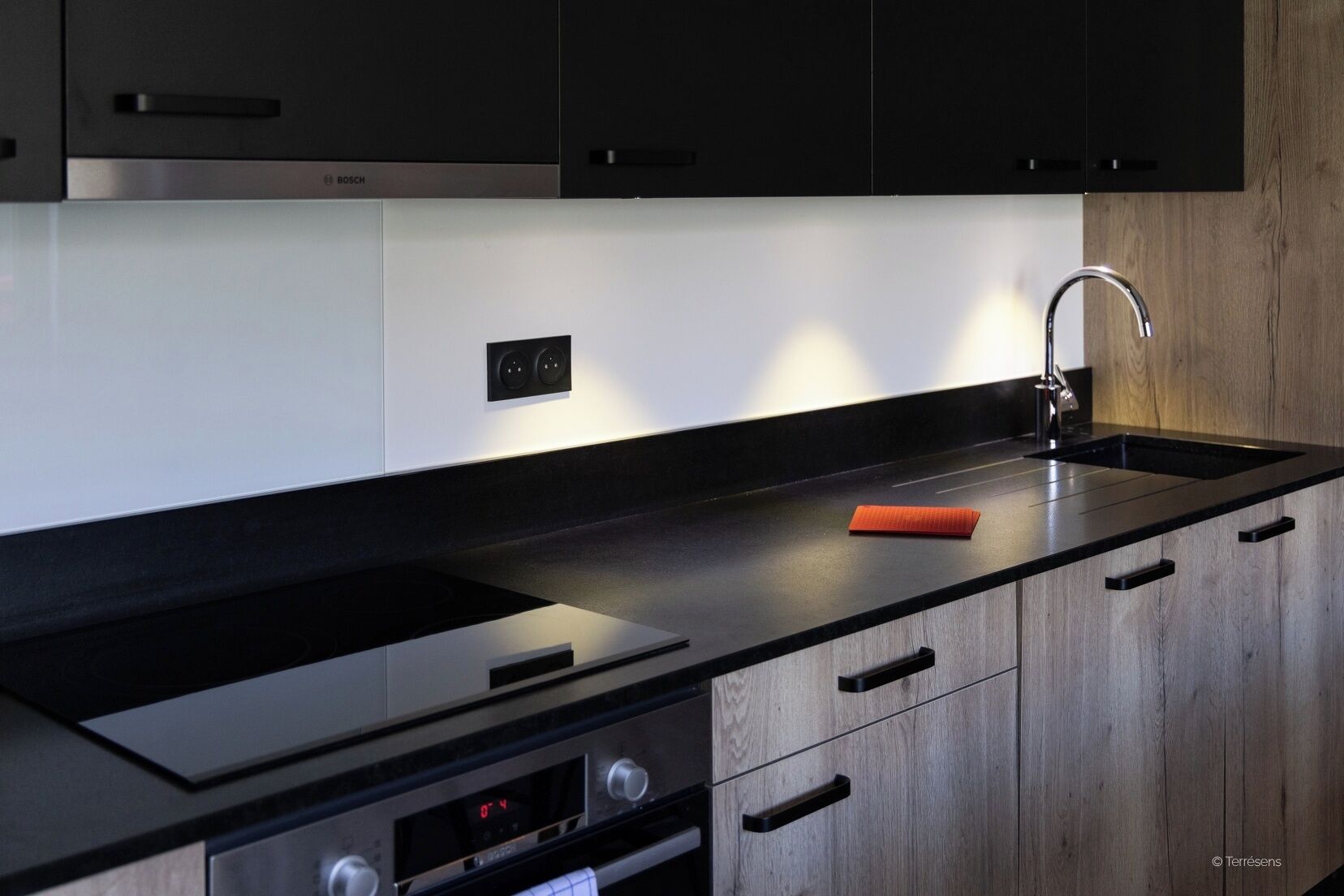 A modern kitchen with black cabinets, a black countertop, a stainless steel range hood, and a built-in oven.