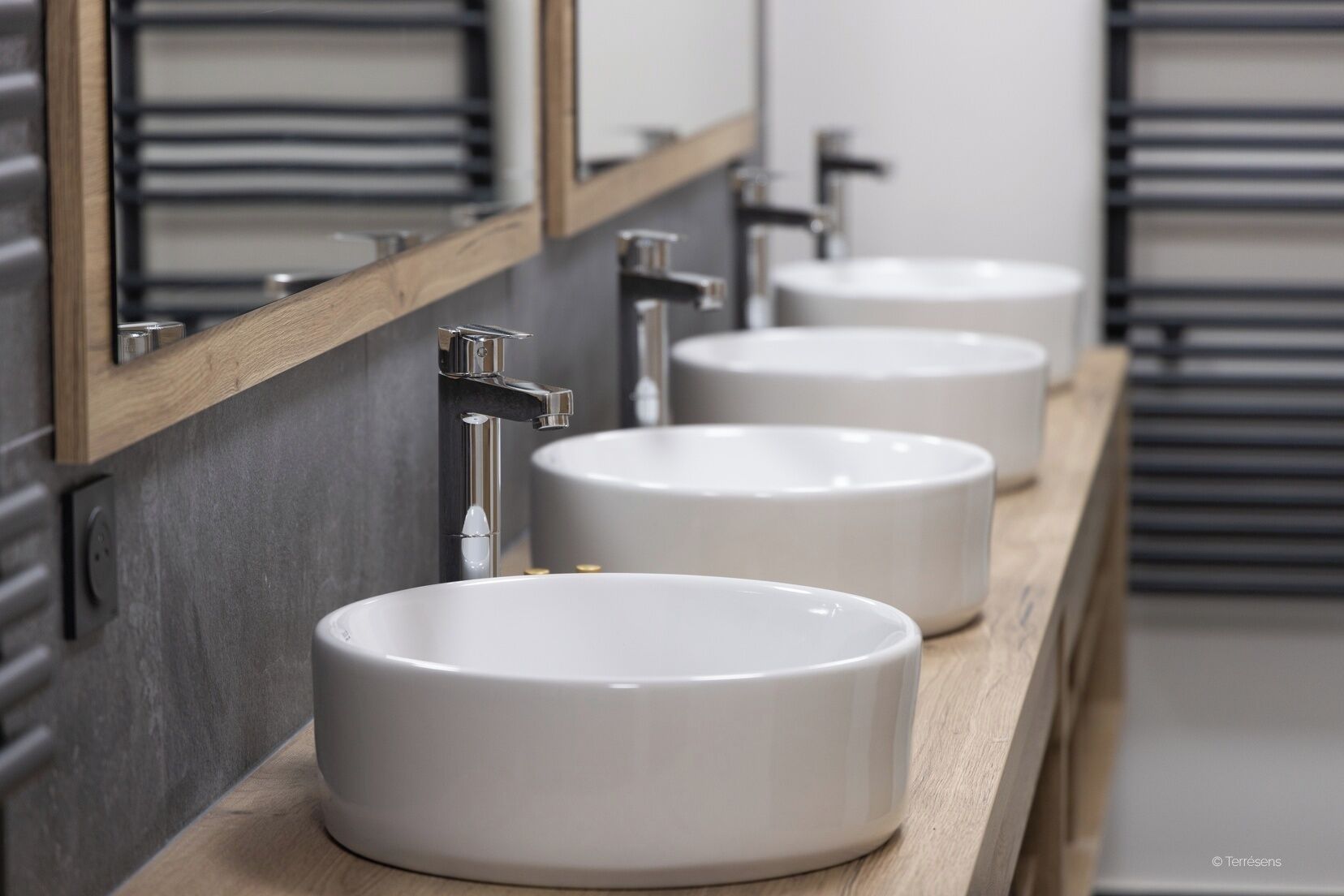 Three white sinks with chrome faucets on a wooden countertop in a modern bathroom.