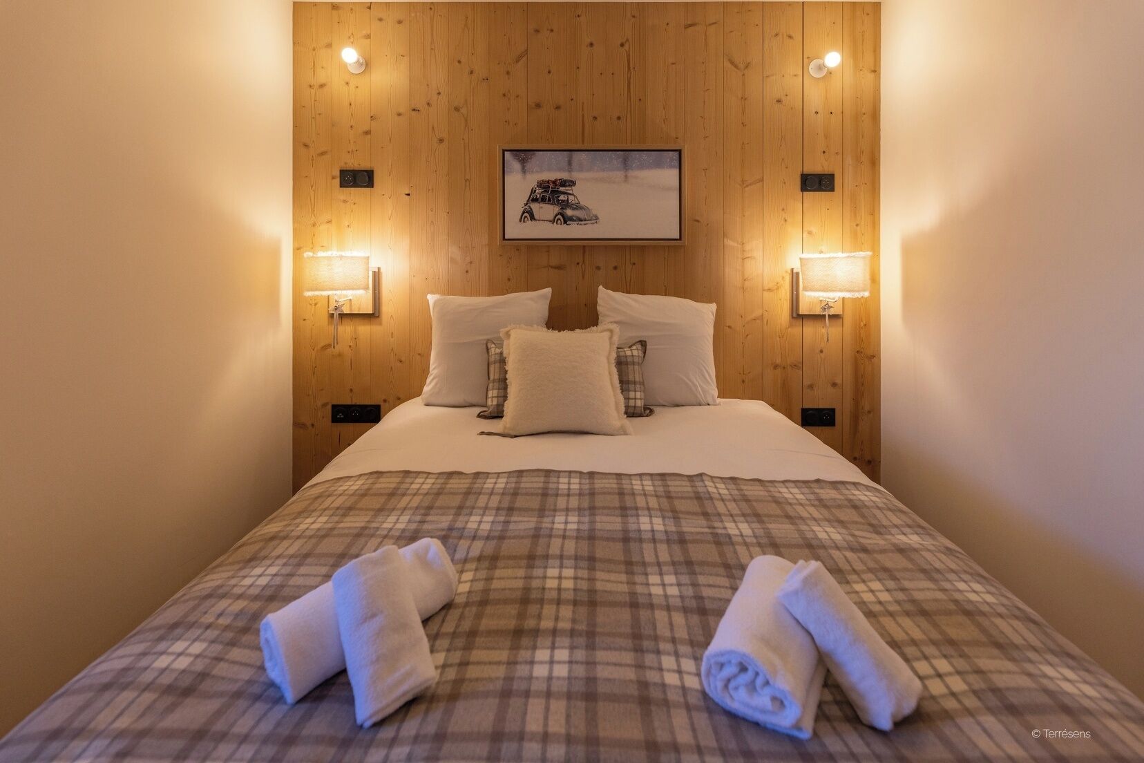 A neatly made bed with plaid bedding, two pillows, and two rolled towels.