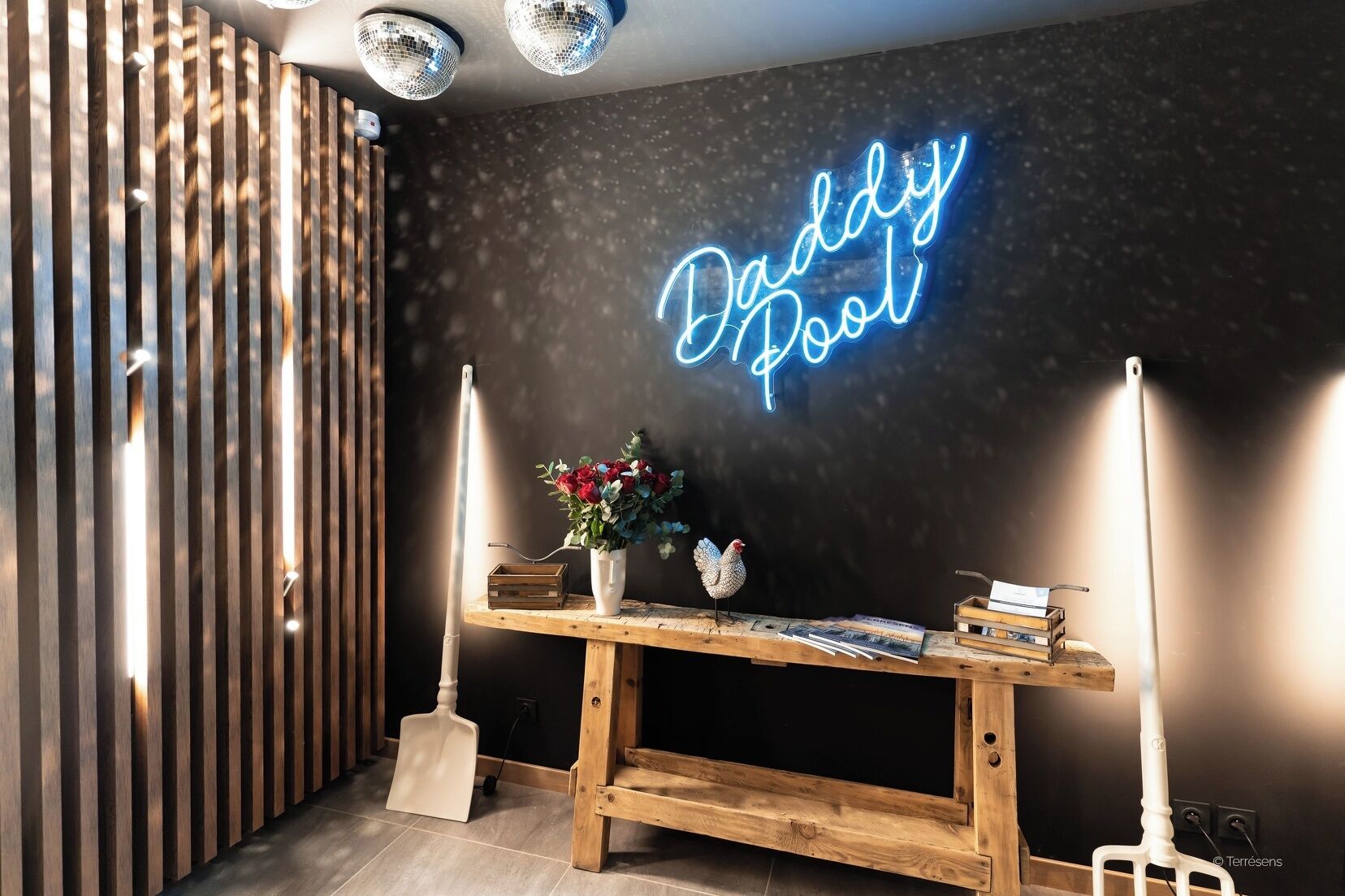A room with a neon sign reading "Daddy Pool," a wooden table with a vase of flowers, books, and a decorative chicken figure.