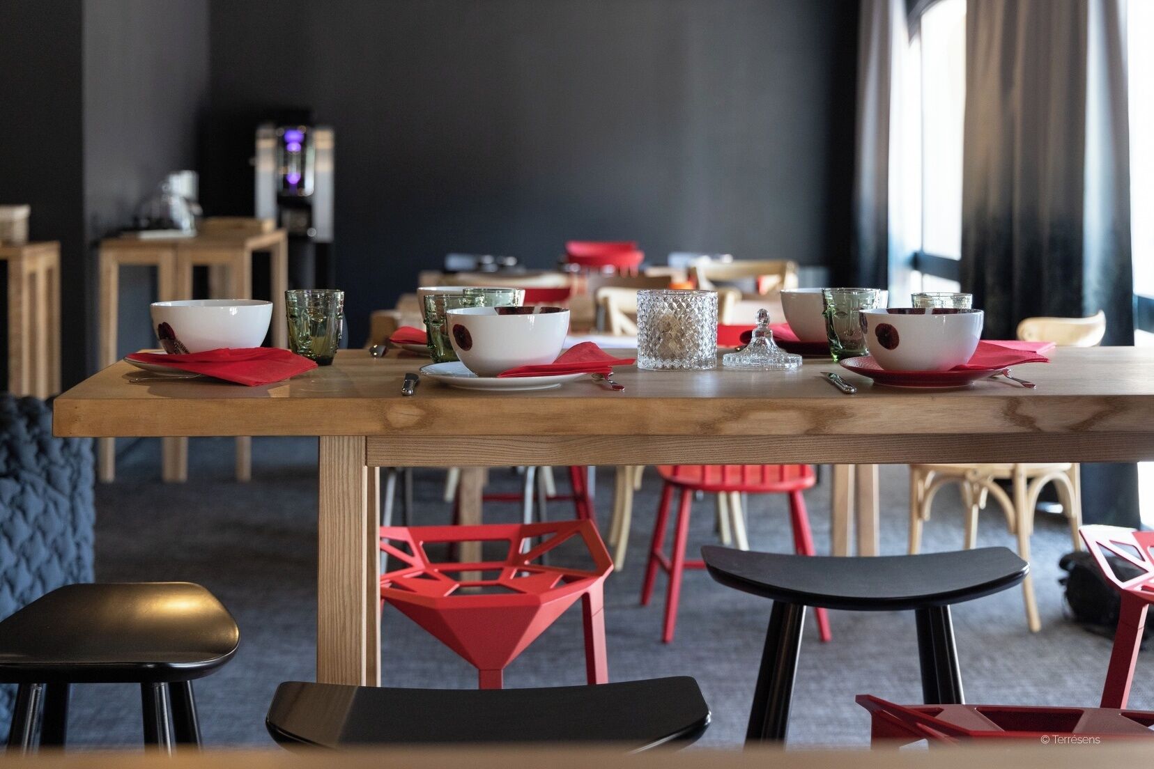 A dining area with a wooden table set for two, red chairs, and a bar area in the background.