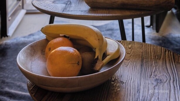 Une table en bois sur laquelle se trouve un saladier rempli de fruits, un plateau avec des tasses et une théière, et, à travers une porte vitrée, on aperçoit un chalet de montagne.
