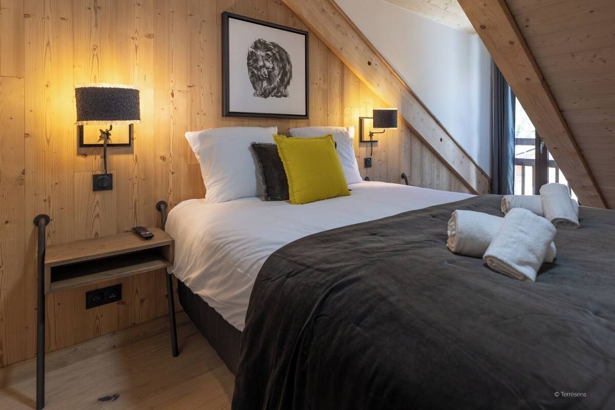 A cozy bedroom with a wooden ceiling, a bed with white and black bedding, a yellow pillow, and two wall-mounted lamps.