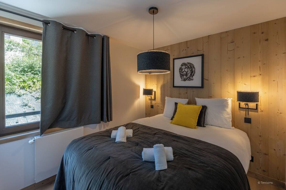 A bedroom with a large bed, a window with curtains, a wall-mounted lamp, and a framed picture.