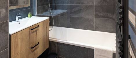Une salle de bain moderne avec des carreaux foncés, un meuble-lavabo en bois, une baignoire blanche et un porte-serviettes noir.
