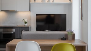 A modern kitchen with a built-in TV, a grey sofa, and a dining area with a wooden table and chairs. - Athenian Riviera Seaside Suite (Vouliagmeni)