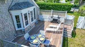A deck with a round table and chairs, a grill, and a small covered area with a window. - Walk to Downtown Chatham, Steps to Beach! (Chatham)