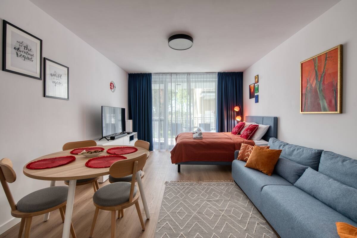 A modern hotel room with a bed, a sofa, a dining table with chairs, a TV, and artwork on the walls.