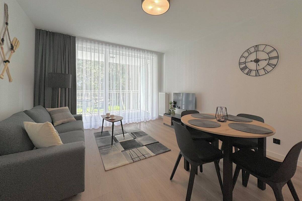 A modern living room with a grey sofa, a dining table with chairs, a large window with sheer curtains, and a wall clock.