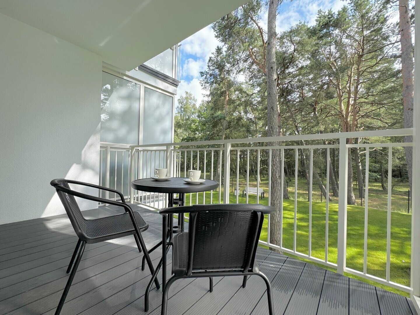 Ein Balkon mit zwei Stühlen und einem kleinen Tisch, der auf eine begrünte Fläche mit Bäumen blickt.