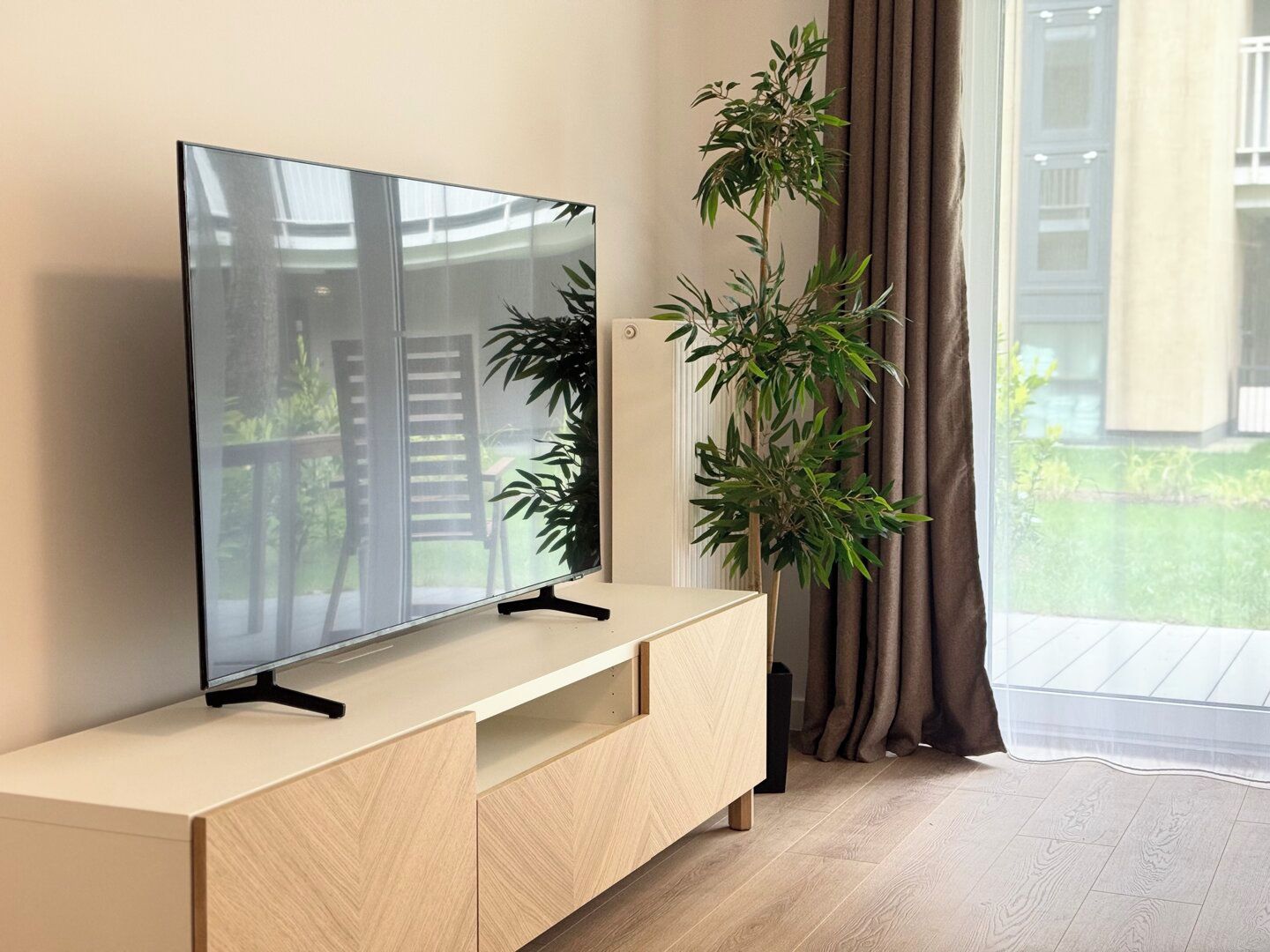 A modern living room with a flat-screen TV on a wooden console, a potted plant, and a view of a balcony.