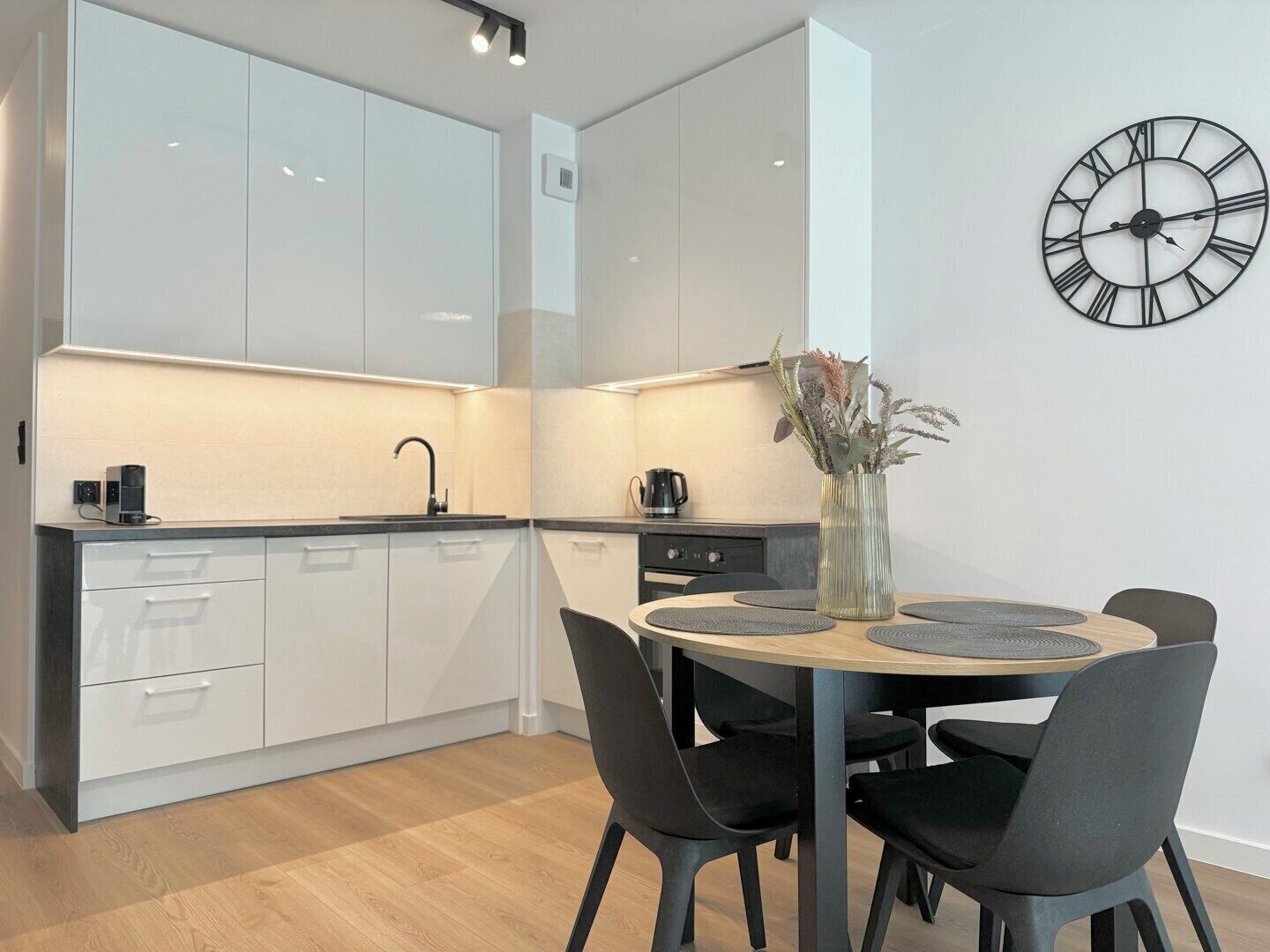 A modern kitchen with white cabinets, a round dining table with black chairs, and a wall clock.