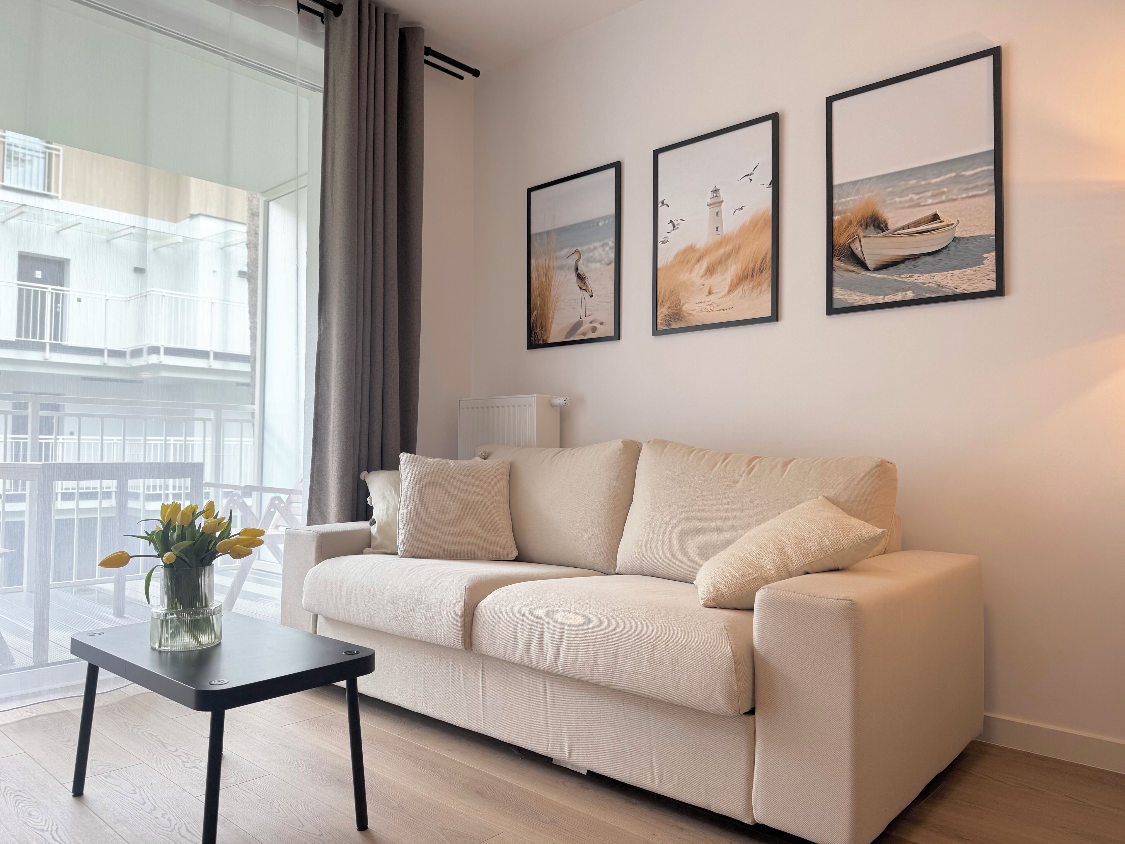 A modern living room with a beige sofa, a small table with a vase of yellow flowers, and three framed pictures on the wall depicting coastal scenes.