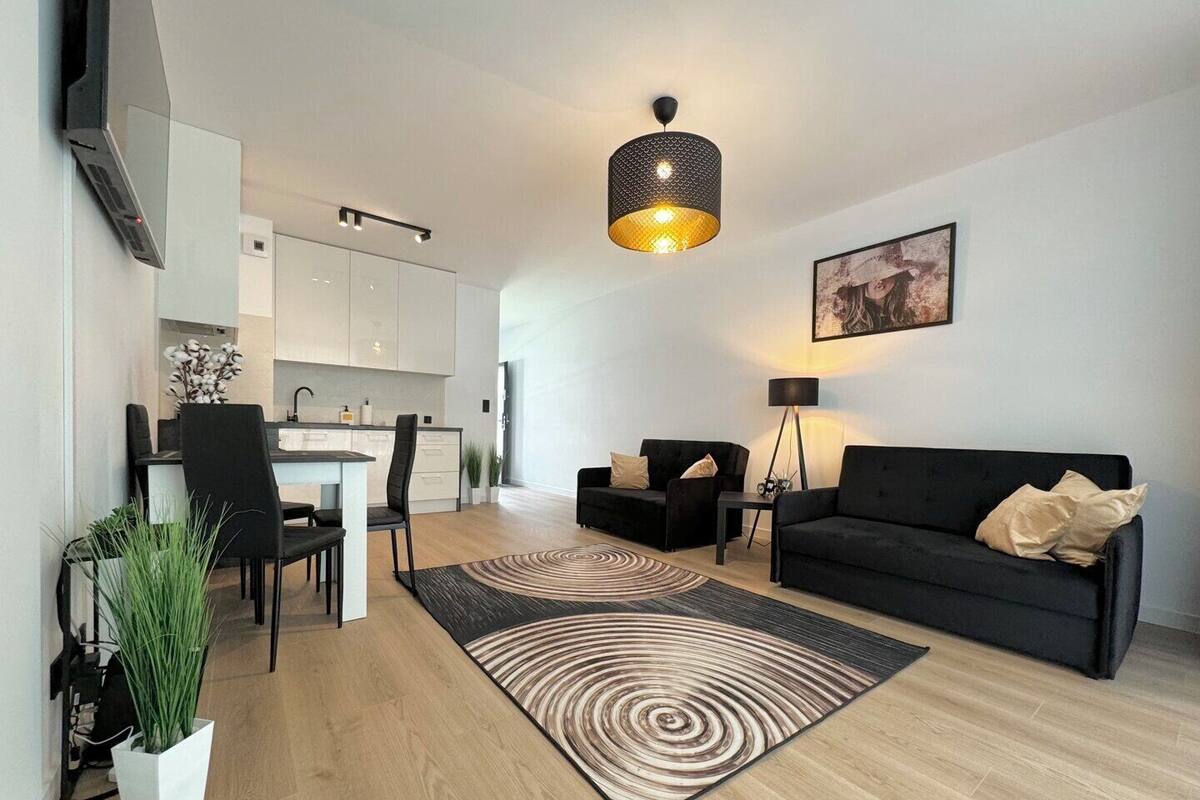 A modern living room with a black sofa, a dining area with a white table, and a kitchen with white cabinets.