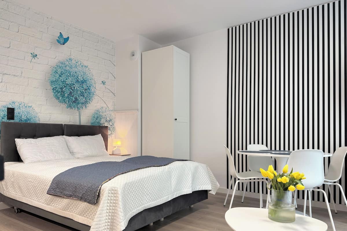 A modern bedroom with a bed, a bedside table with a lamp, a wall with a blue tree mural, a striped wall, and a small round table with a vase of flowers.