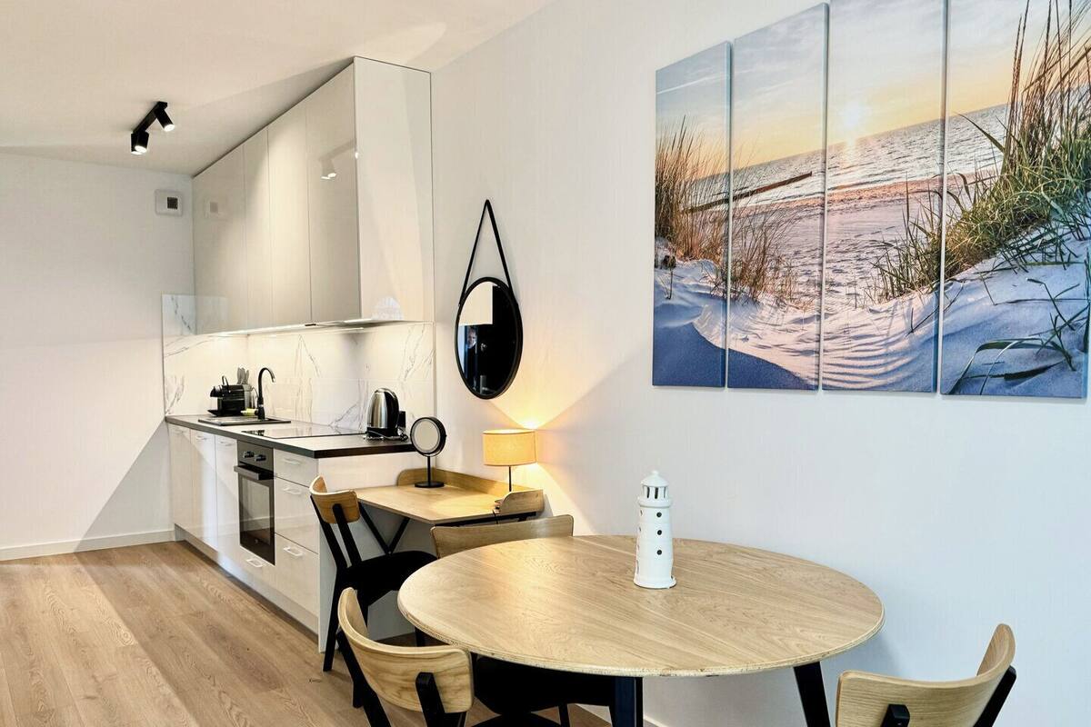 A modern kitchen with white cabinets, a wooden dining table, and a painting of a snowy beach scene.