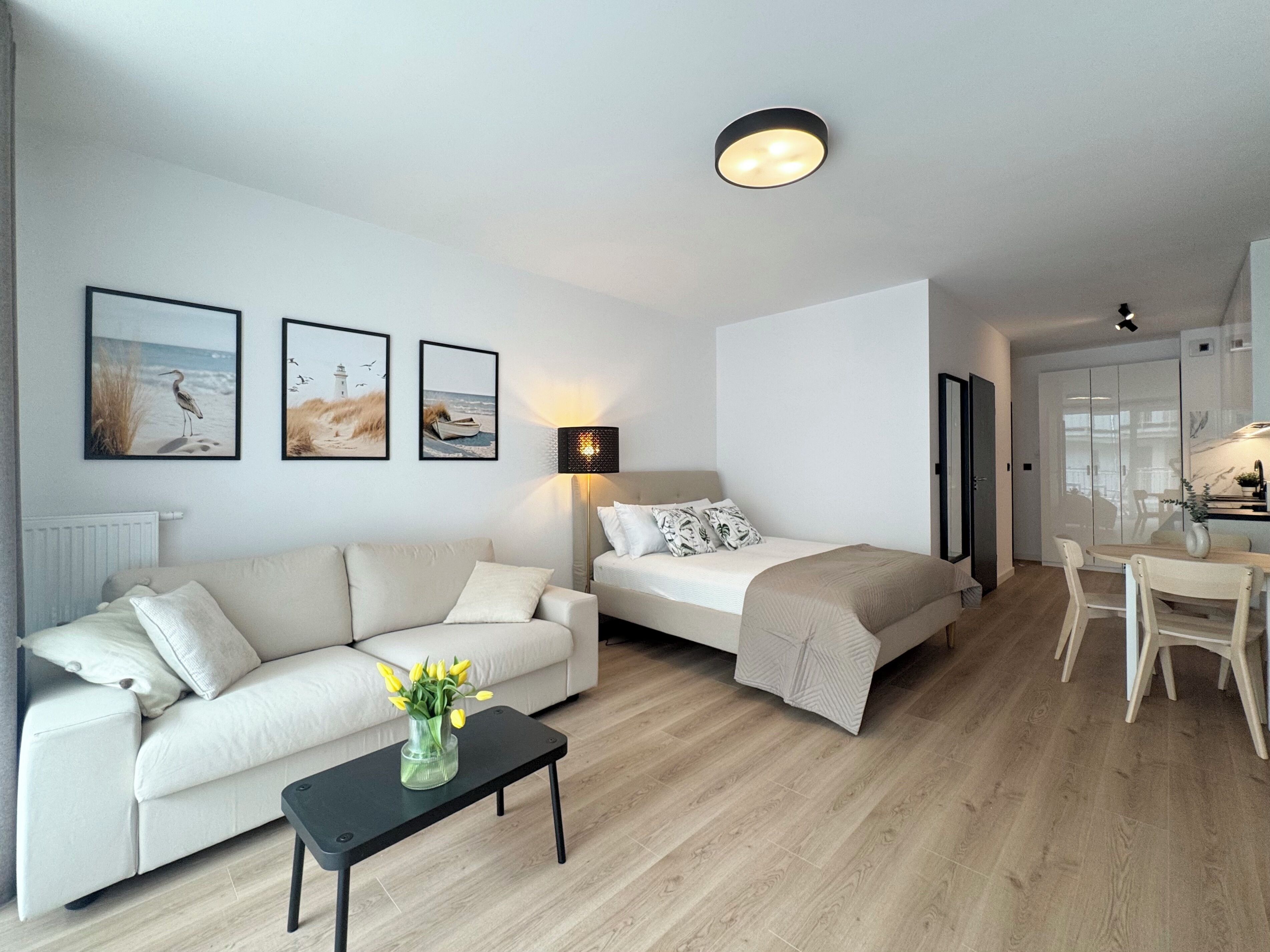 Een moderne, goed verlichte woonkamer met een bank, een bijzettafeltje met een bloevas, een bed met hoofdbord, en een eethoek met tafel en stoelen.