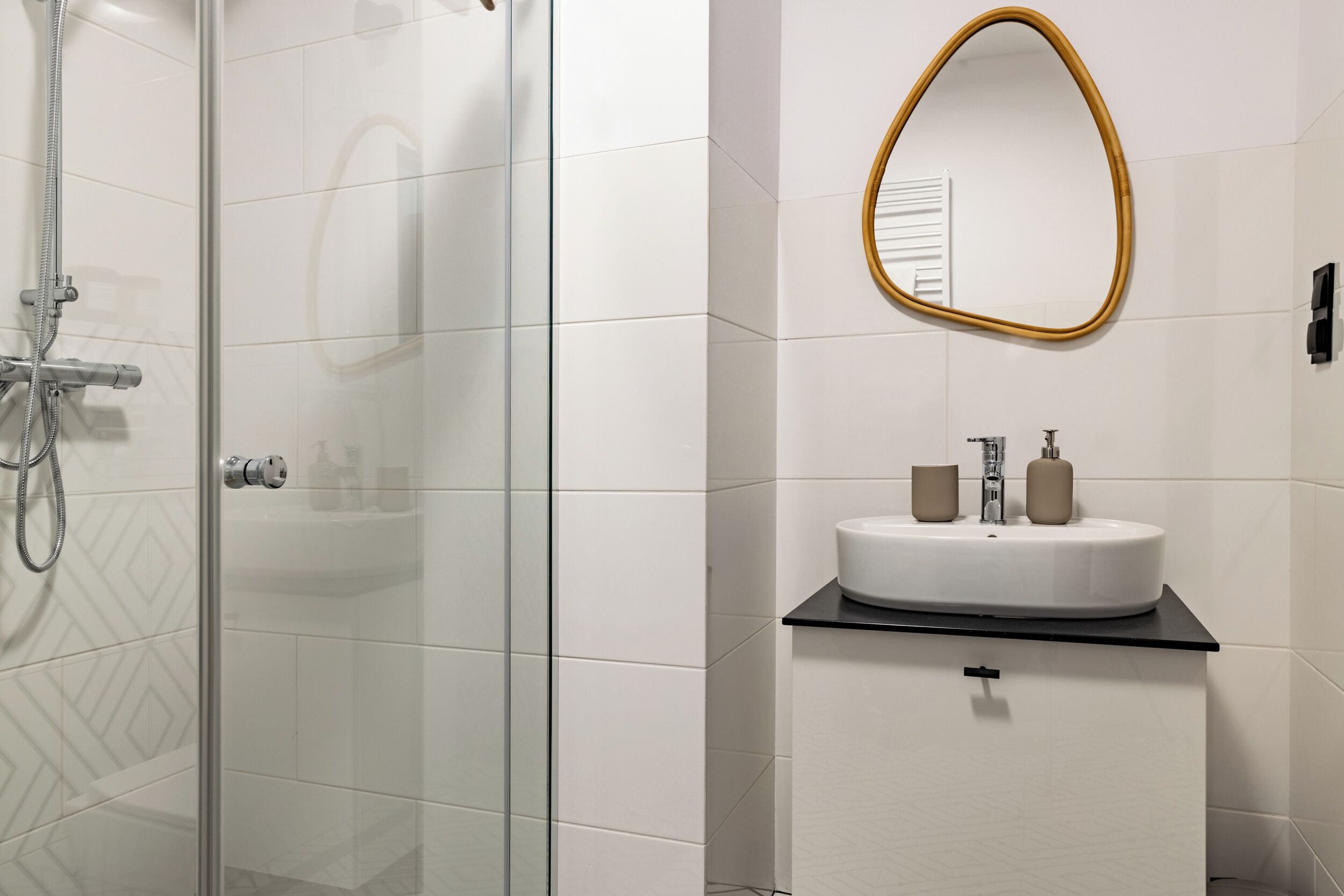 Une salle de bain moderne avec une douche à l’italienne en verre, un lavabo blanc doté de deux distributeurs de savon et un miroir avec un cadre doré.