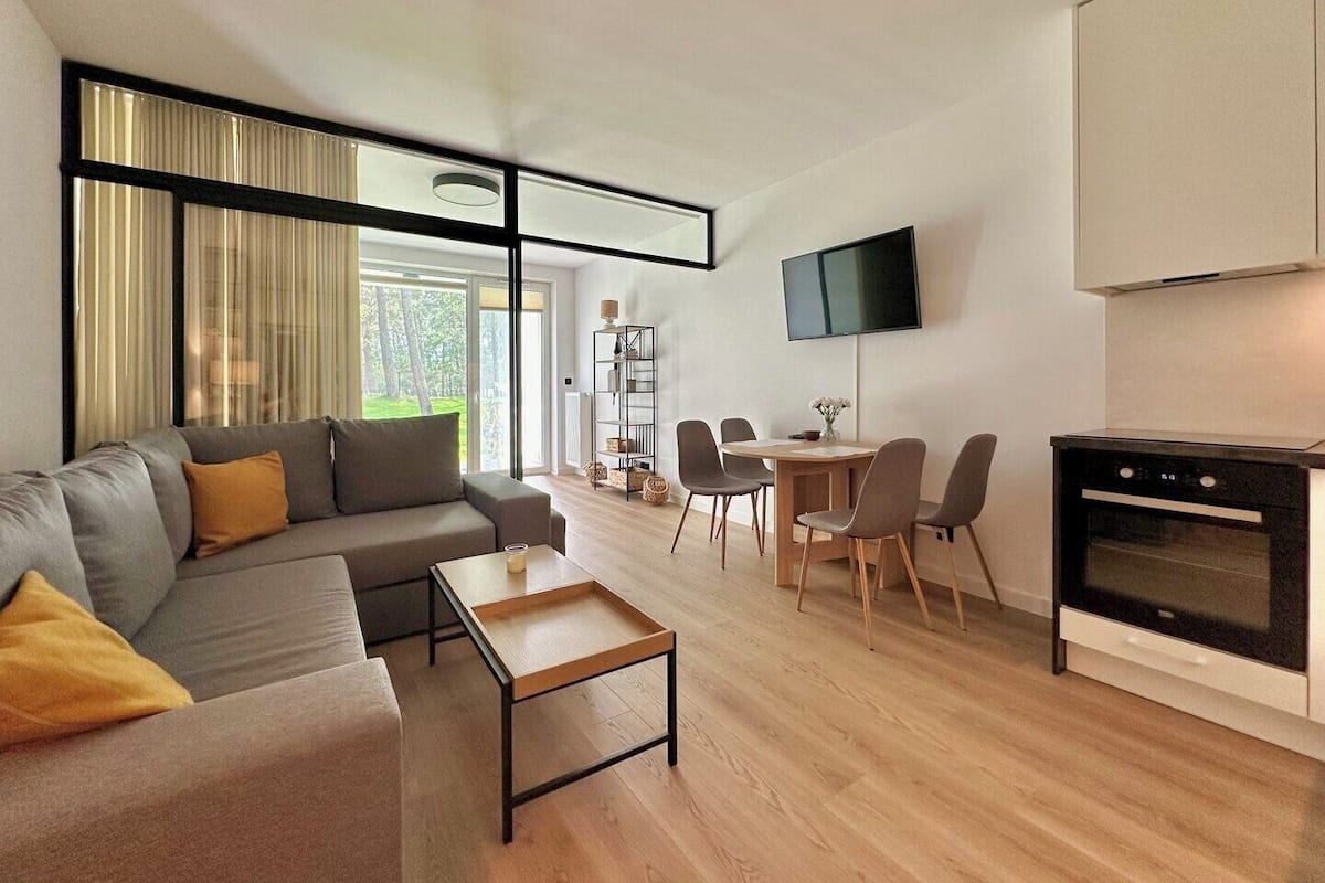 A modern living room with a sofa, coffee table, and dining area with chairs. There is a flat-screen TV mounted on the wall and a kitchen area with an oven.