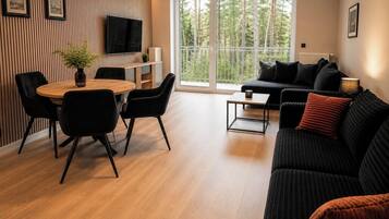 Una sala de estar moderna con una mesa de comedor de madera, sillas negras, un sofá negro con cojines rojos, un televisor montado en la pared y un gran ventanal con vistas a árboles.