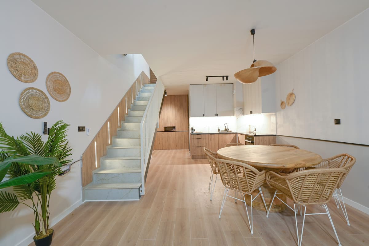 A modern interior with a staircase, a dining area with a round table and wicker chairs, and a kitchen with wooden cabinets and a hanging light fixture.