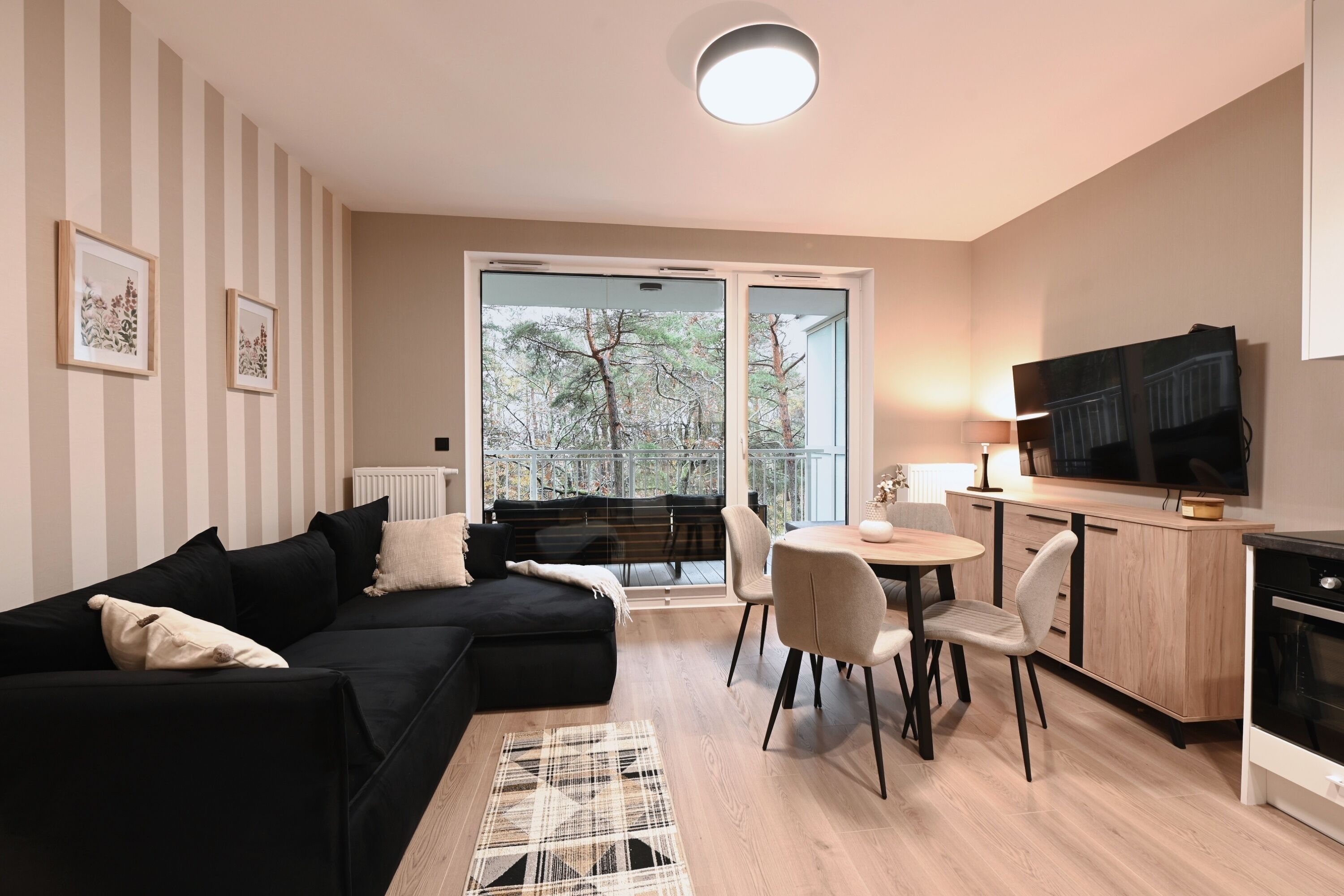 A modern living room with a black sofa, a dining table with chairs, a TV, and a kitchen area.