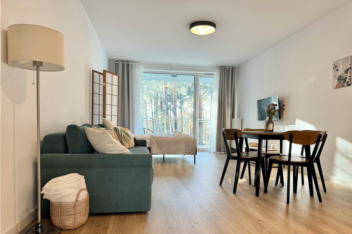 Una sala moderna con sofá, una mesa de comedor con sillas, una lámpara de pie y un gran ventanal con cortinas.
