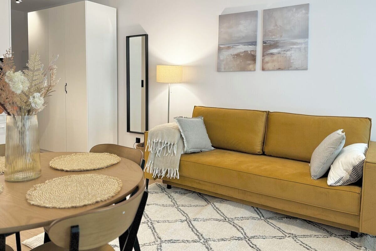 A modern living room with a wooden dining table, a mustard-colored sofa, and a patterned rug.