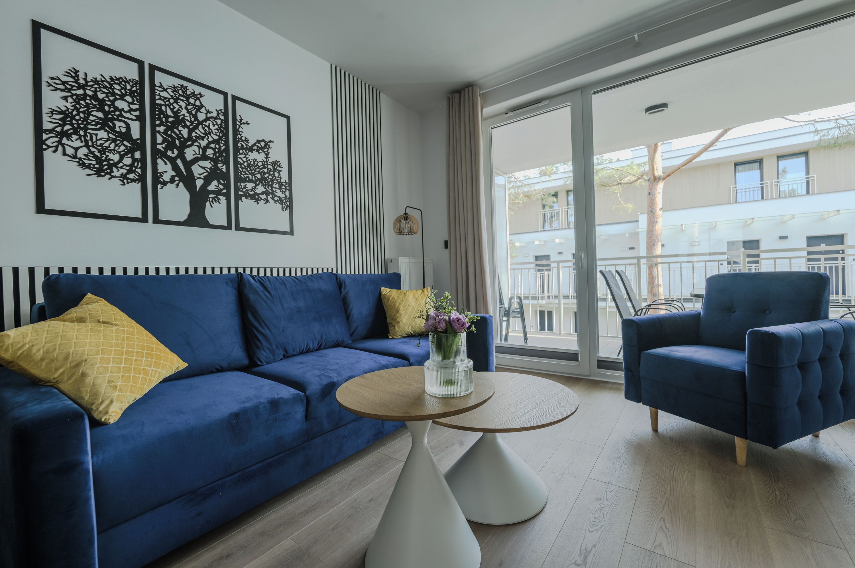 A modern living room with a blue sofa, a matching armchair, and a round coffee table. There are three framed artworks on the wall and a window with a view of the outside.