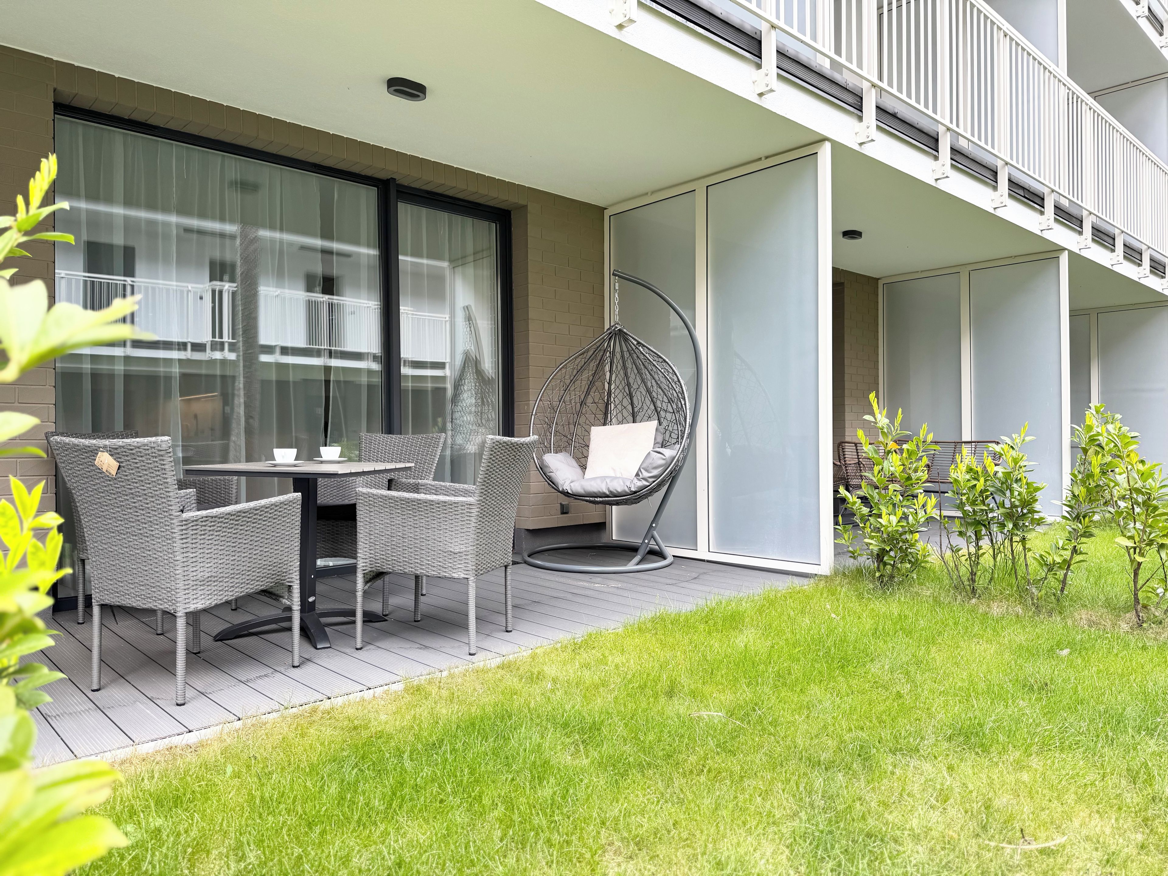 A modern outdoor seating area with wicker furniture and a hanging chair, adjacent to a building with large windows and a glass door.