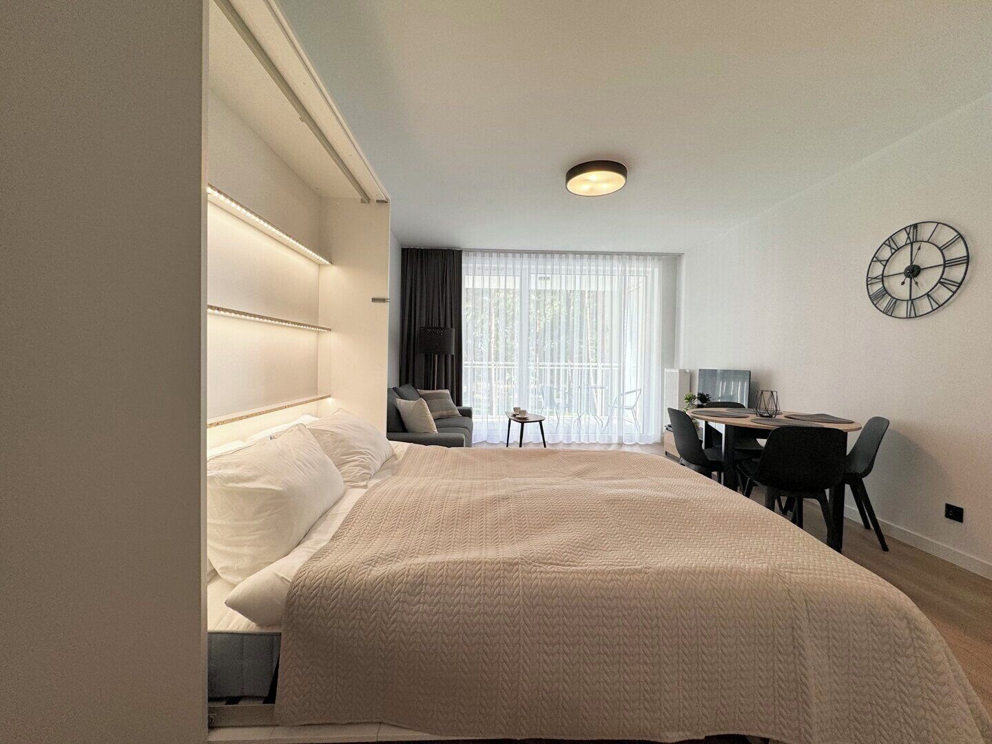 A modern hotel room with a large bed, a desk with a chair, a small table, and a large window with curtains.