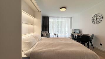 A modern hotel room with a large bed, a desk with a chair, a small table, and a large window with curtains.