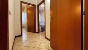 A hallway with a wooden door on the right, a door at the end, and a bed with a blue and white checkered bedspread in the background. - Comfortable flat in Bibione - Beahost (Rosolina Mare)