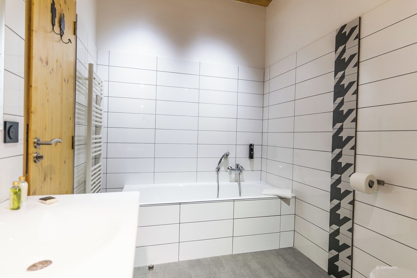 A modern bathroom with white tiles, a bathtub, a wooden door, and a towel rack.