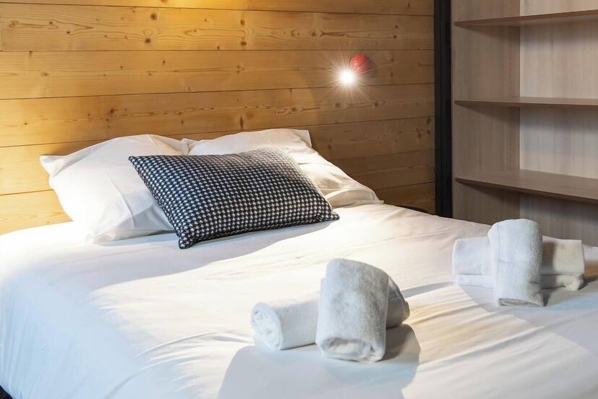 Een net opgemaakt bed met witte lakens, een gestreept kussen en opgerolde handdoeken op een houten vloer.