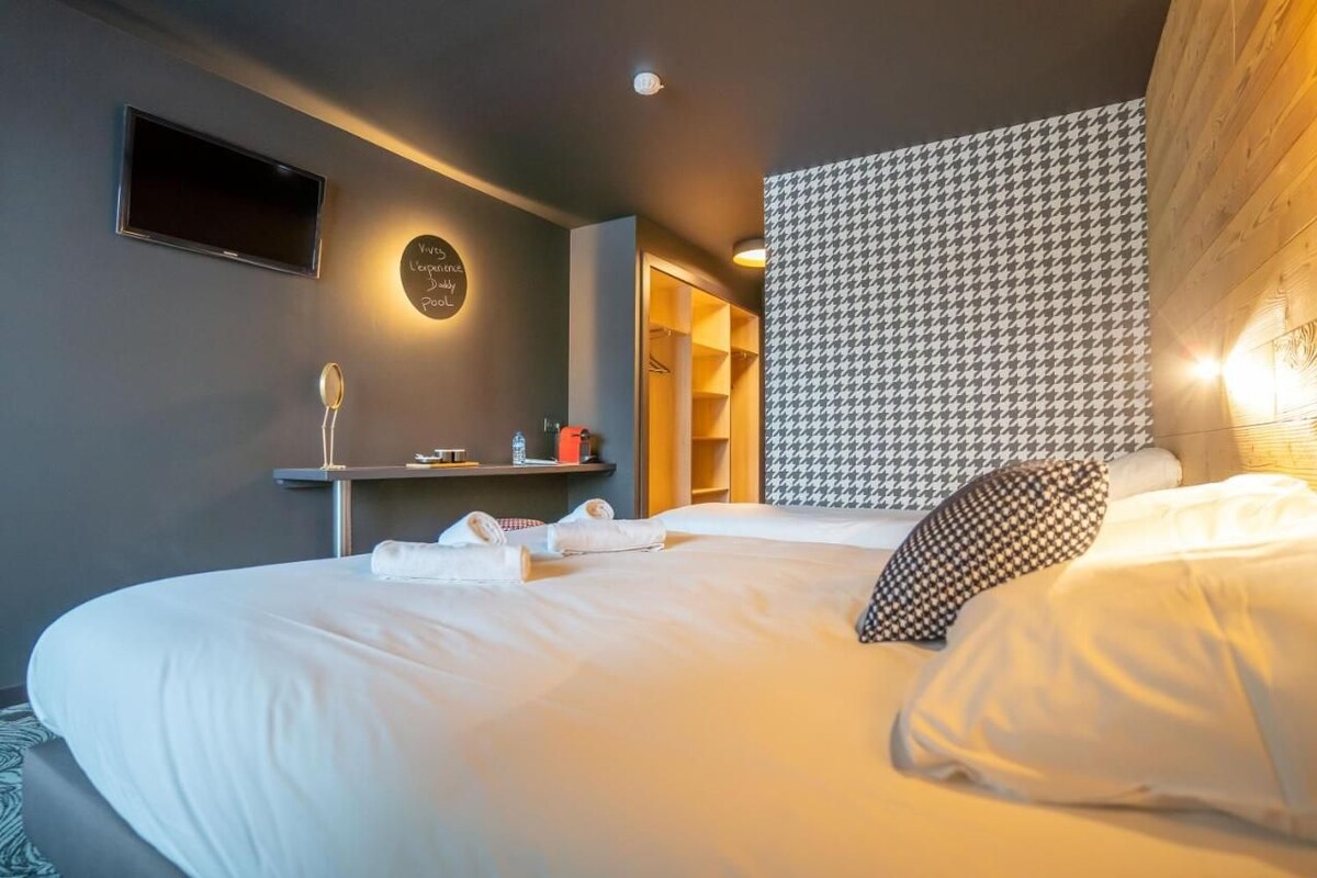Une chambre d’hôtel moderne avec un grand lit, un téléviseur fixé au mur, une table de chevet avec une lampe et une tête de lit à motifs.