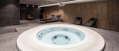 A modern indoor swimming pool with a hot tub, lounge chairs, and a tiled floor.