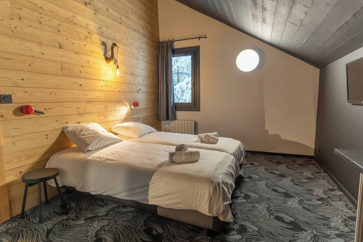 Een kamer met twee bedden, een houten wand, een raam met gordijnen, een ronde lamp en een radiator.