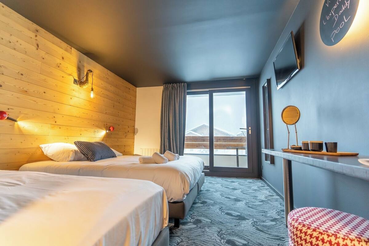 Een hotelkamer met twee bedden, een houten wand, een groot raam en een klein tafeltje met kopjes en een spiegel.