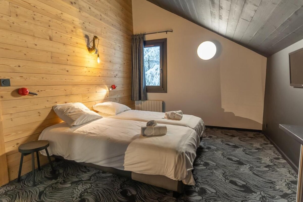 Ein gemütliches Zimmer mit zwei Betten, einer runden Deckenleuchte, einem Fenster mit Blick auf Bäume und einer Holzwand.